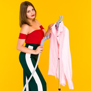 Young woman steaming pink shirt at dry-cleaner's on yellow background