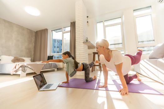 Mother and daughter practicing online yoga classes at home during the quarantine during the coronavirus pandemic. Family doing sports together online from home during isolation together at home