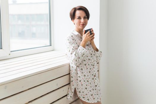 Beautiful young woman with cup of tea standing near the window at home
