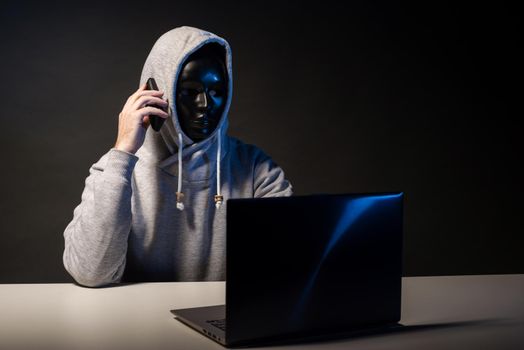 Anonymous hacker in mask programmer uses a laptop and talking on the phone to hack the system in the dark. The concept of cybercrime and hacking database