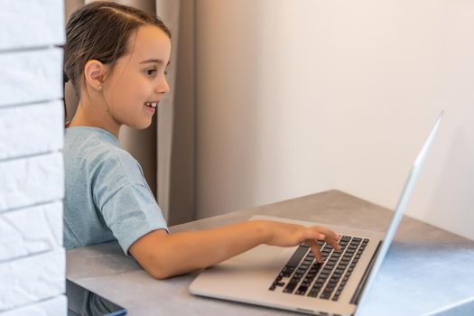 Little girl of school age is studying at home. A girl watches a video lesson on a laptop monitor and writes tasks in a notebook.