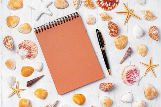 Open notepad with sea shells on white wooden background, top view