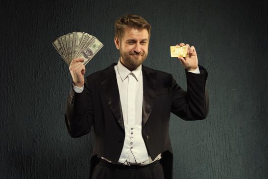 Positive man in a tailcoat offers a credit card and money. - image