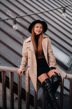 A stylish young woman in a beige coat and black hat sits on a rooftop in the city center. Women's street fashion. Autumn clothing.Urban style.