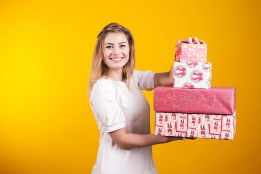 Portrait of a happy smiling girl in dress shows present boxes isolated over yellow background.