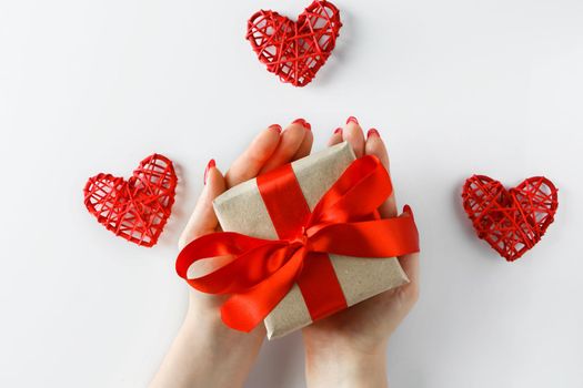 Gift with a red ribbon in hands on a white background. Giving a gift for Valentine's Day. Box with a satin ribbon in the hands of a woman. Red hearts on a white background.