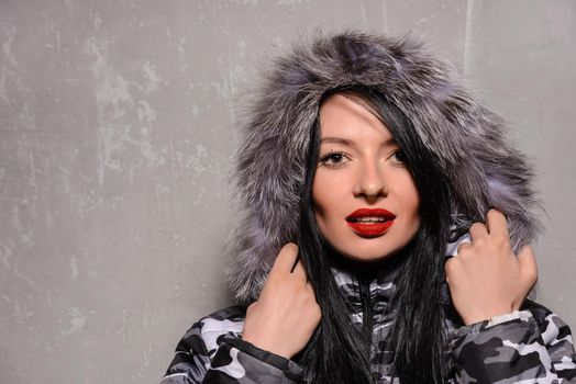 Sweet young woman in warm winter jacket with fur hood isolated on grey background.