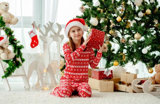 Pretty child girl holding Christmas red gift box in decorated room. Kid celebrating New Year with presents at home