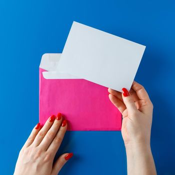 Pink envelope with a blank card in hands on a blue background. Pink envelope with a greeting card in the hands of a young woman.