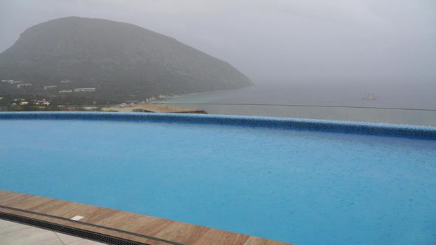 Autumn rain falls in large drops on the heated outdoor pool. Swimming pool in the hotel on the background of the mountain and the sea