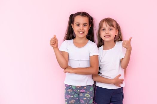 Children showing like. Two adorable happy little girls gesturing thumbs up together and smiling to camera, excellent feedback, good job. indoor studio shot isolated on pink background