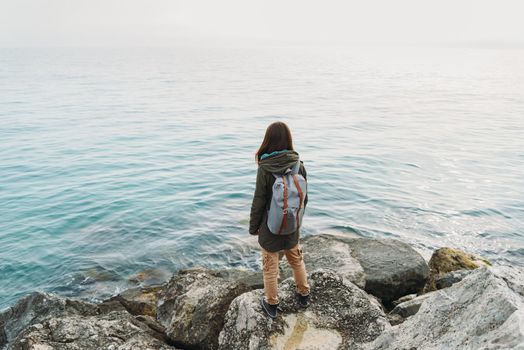 Traveler young woman standing on stone on coastline and enjoying view of sea, rear view