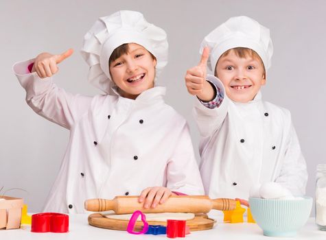two smiling children rolloing out a dough in the kitchen with thumbs up