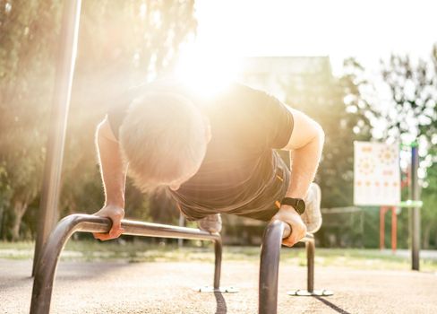 Strong man guy training and doing push ups with horizontal bar with sunlight at the stadium outdoors. Adult male person during workout for arms and chest. Sportsman exercising outside
