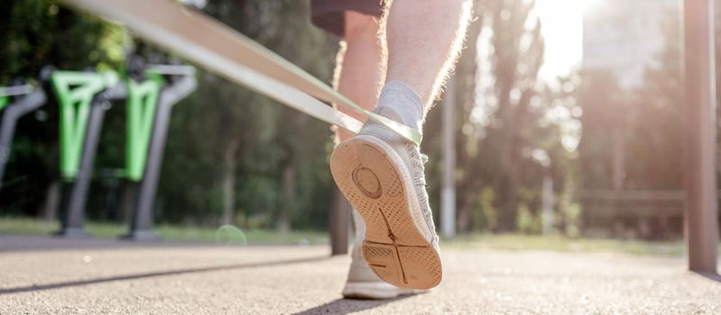 Closeup back view of legs feets of man guy training with elastic rubber band at the stadium outdoors. Adult male person during workout with additional sport equipmant with sunlight