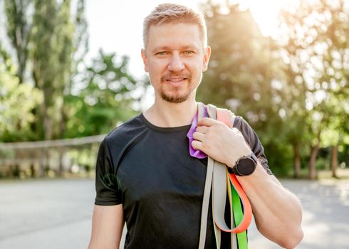 Sportive man guy holding in his hand set of colorful elastic rubber bands and smiling looking at the camera at the stadium outdoors. Athlete male person during workout with additional sport equipmant