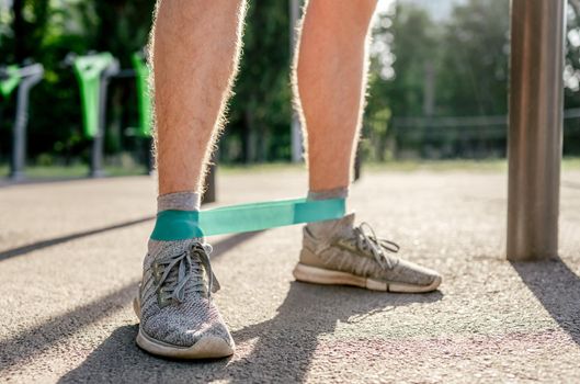 Closeup front view of legs feets of man guy training with elastic rubber band at the stadium outdoors. Athlete adult male person during active workout with additional sport equipmant in summertime