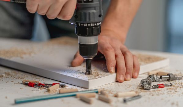 Man working using electric screwdriver during process of wooden furniture manufacturing in workshop
