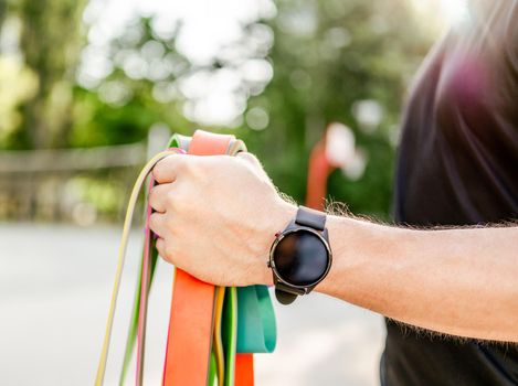 Strong man guy holding in his hand wearing fitness tracker set of colorful elastic rubber bands at the stadium outdoors. Athlete male person during workout with additional sport equipmant