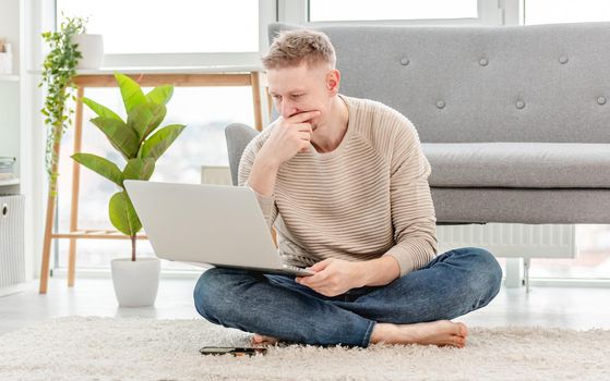 Man freelancer looking at laptop and thinking during remote work time at home. Adult guy sitting on the floor searching in internet