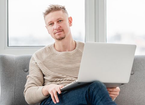 Attractive blond man freelancer sitting on the sofa holding laptop. Remote work at home