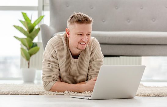 Man searching and reading from laptop lies on the floor at home in sunny day. Freelance remote job with modern technologies