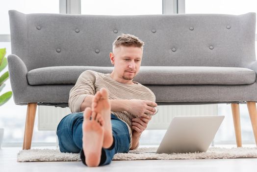 Man sitting on the floor with coffee and reading in laptop at home. Light beige interior workspace