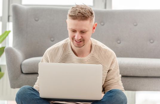 Attractive blond man sitting with laptop, typing and chatting using internet. Home workspace during remote daily work