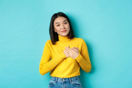 Portrait of beautiful heartfelt woman holding hands on heart, smiling and listening compassionate, standing over blue background in yellow pullover.