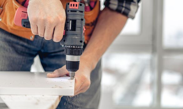 Man working using electric screwdriver during process of wooden furniture manufacturing in workshop