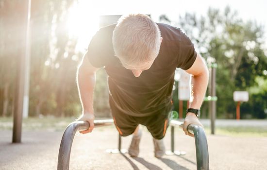Strong man guy training and doing push ups with horizontal bar with sunlight at the stadium outdoors. Male person during workout for arms and chest. Sportsman exercising