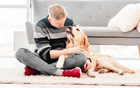 Man sitting with golden retriever dog and hugging it on the floor. Doggy kissed owner