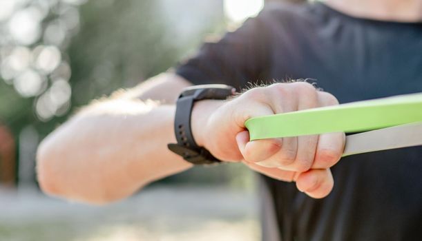 Closeup view of hands of man guy training with elastic rubber band at the stadium outdoors. Adult male person during workout for arms and chest with additional sport equipmant