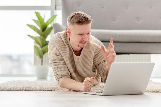 Adult blond man freelancer lies on carpet with laptop and captured at the work moment. Guy thinking during remote working day