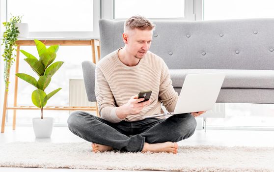 Man freelancer working at home sitting with laptop and smartphone on the floor. Remote work in Covid quarantine time