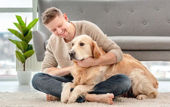 Man sitting on the floor and hugging golden retriever dog. Owner petting his doggy in sunny room at home