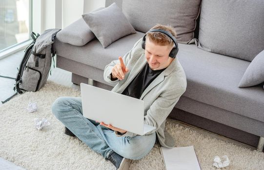 Cheerful man talking online on computer sitting on floor in light apartment