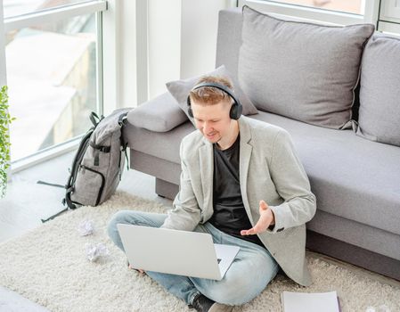 Cheerful man talking online on computer sitting on floor in light apartment