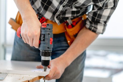 Man working using electric screwdriver during process of wooden furniture manufacturing in workshop