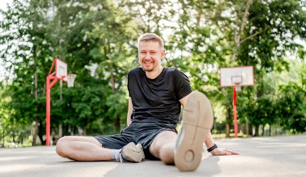 Sportive man guy sitting at the stadium outdoors, smiling and looking at the camera. Portrait of athlete male person during workout outdoors