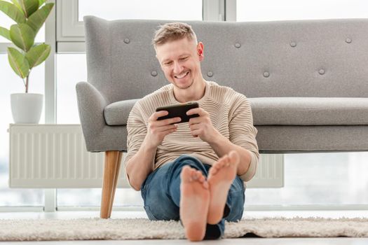 Attractive blong man playing games on smartphone, sitting on the floor and smiling. Online global gaming connection