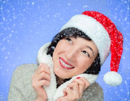 Cute young woman in santa hat is smiling in snowy weather on blue background
