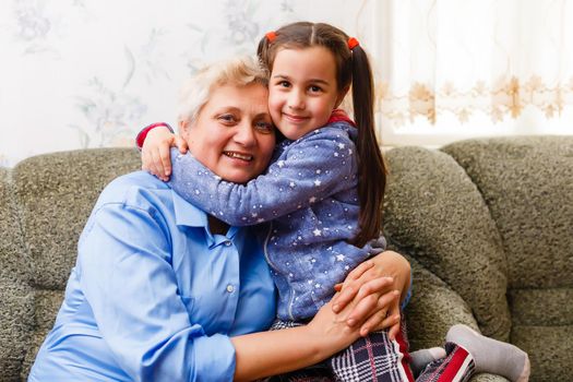 Little cute girl and her grandmother are spending time together at home. Having fun, hugging and smiling while sitting on sofa.