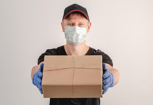 Young courier in protective mask and gloves holding small parcel on white wall background