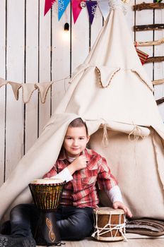boy sitting on the floor and playing drum near wigwam in decorated wooden room