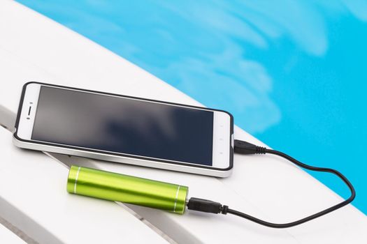 Smartphone connected to the green portable battery charger through USB cable on blue water background. Emergency charging of the phone on a lounger near the swimming pool during the rest.