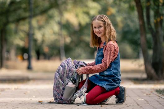 Preteen school girl with backpack sitting on the ground and smiling. Pretty pupil female kid with schoolbag in autumn park