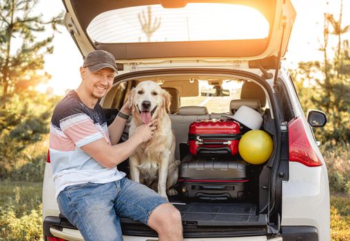 Man with golden retriever sitting in open car trunk next to luggage