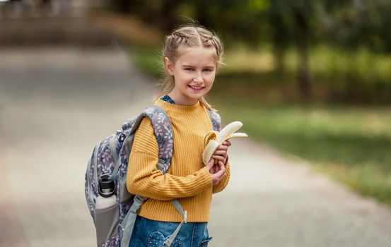 Preteen school girl with backpack holding bananas in her hands, looking at camera and smiling. Pretty pupil female kid after education class