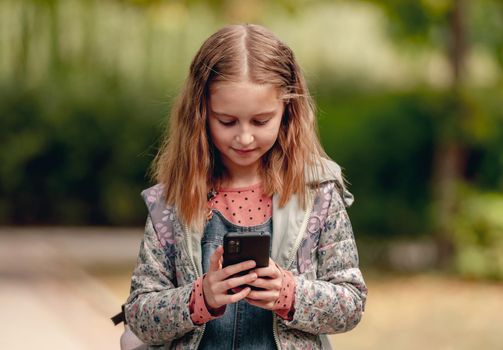 Pretty school girl with smartphone outdoors. Cute female kid standing with cell phone in autumn park